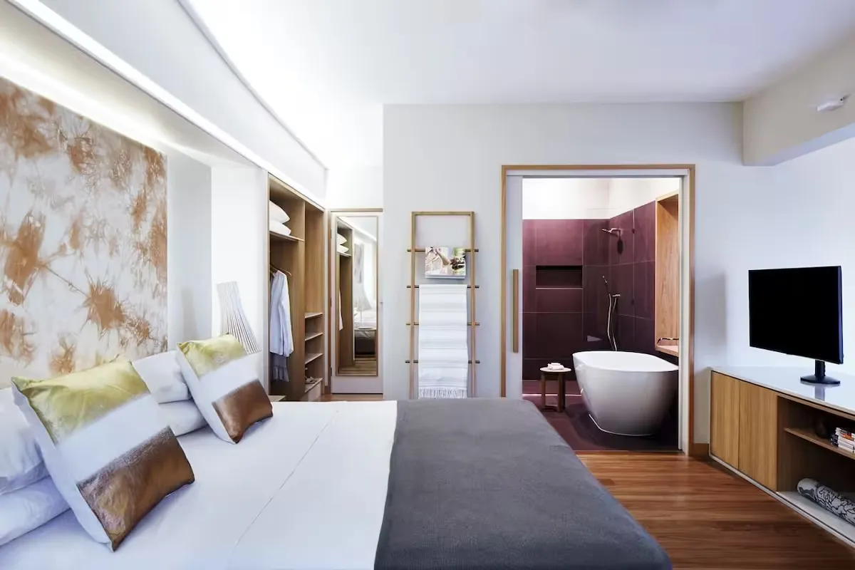 Hotel suite with a tie-dyed art wall behind the bed, an open wardrobe and a freestanding bath in the ensuite