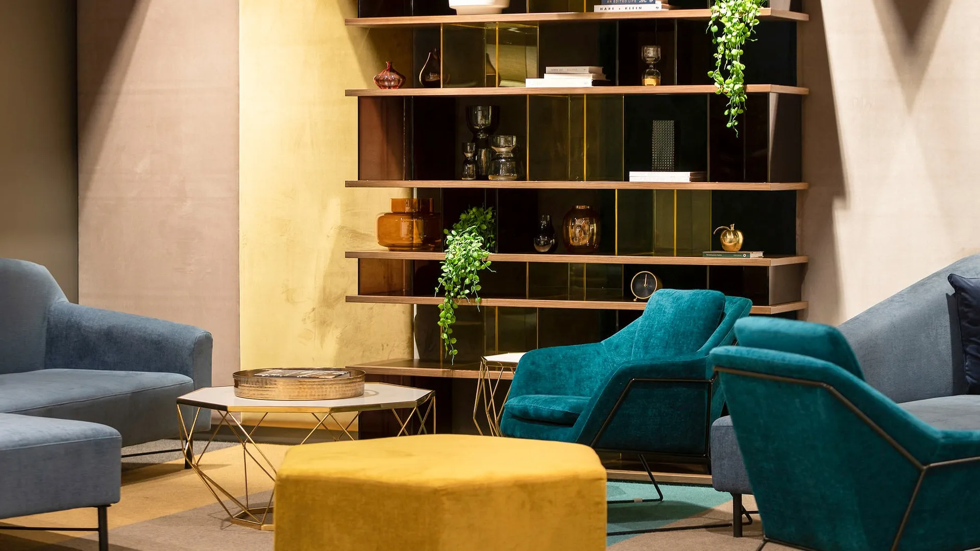 Teal armchairs and a mustard ottoman beside a brass and timber shelving wall
